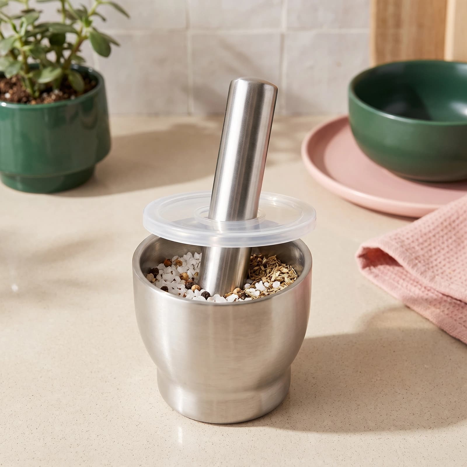 Silver brushed stainless steel mortar and pestle set with a clear plastic lid, filled with spices, on a light kitchen counte…