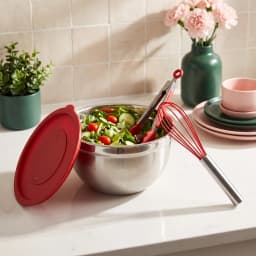 Stainless steel mixing bowl with red whisk and red lid on a light kitchen counter, next to a pink potted plant. - view 7
