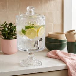 Clear glass beverage dispenser with decorative patterned body, silver spigot, and matching lid on a white background. - view 1