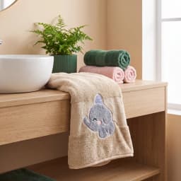 Folded beige microfiber towel with a grey elephant wearing a blue striped hat embroidered on it, on a grey textured surface. - view 1