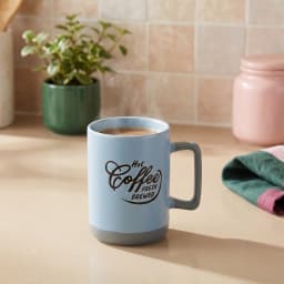 كوب سيراميك أزرق فاتح بقاعدة ويد رماديين، عليه كتابة "Hot Coffee Fresh Brewed" وبخار طالع منه، على رخامة مطبخ. - view 1