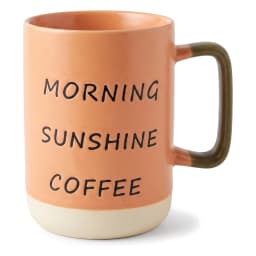 كوب سيراميك برتقالي بعبارة 'MORNING SUNSHINE COFFEE' بالأسود، يد باللون الزيتوني وقاعدة كريمي على رف أبيض. - view 2