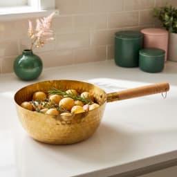 Golden hammered stainless steel pan with a light wooden handle, filled with small potatoes, rosemary, and garlic on a white… - view 1