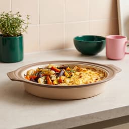 Beige and cream oval ceramic baking dish filled with roasted vegetables and a cheesy gratin, on a light kitchen counter. - view 1