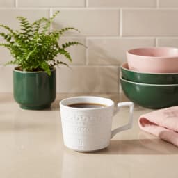 Cream ceramic mug with a textured pattern and square handle, filled with coffee, on a light countertop with a plant and bowl… - view 1