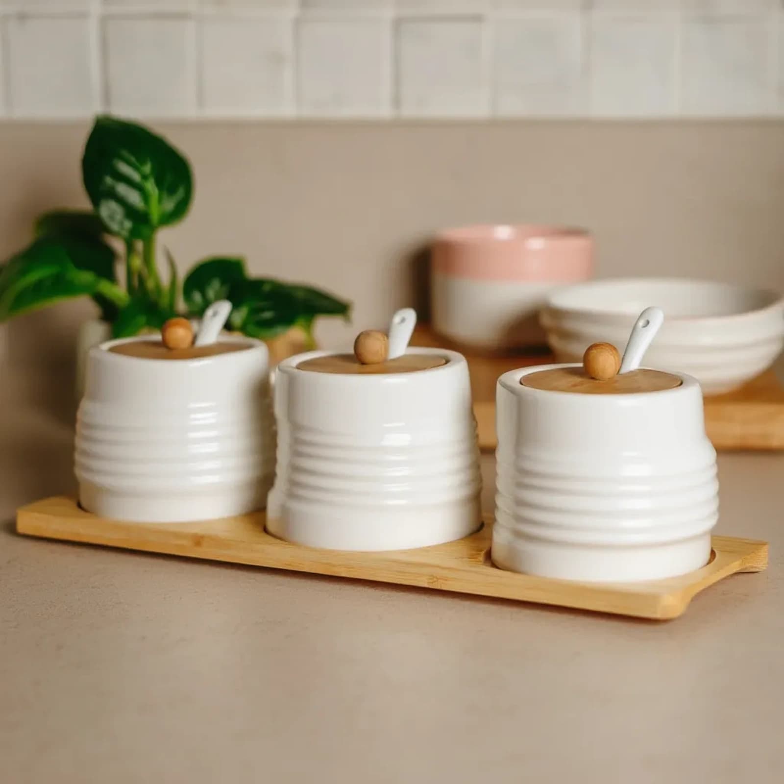 Three white ribbed porcelain jars with light brown wooden lids and small white ceramic spoons on a rectangular bamboo tray.