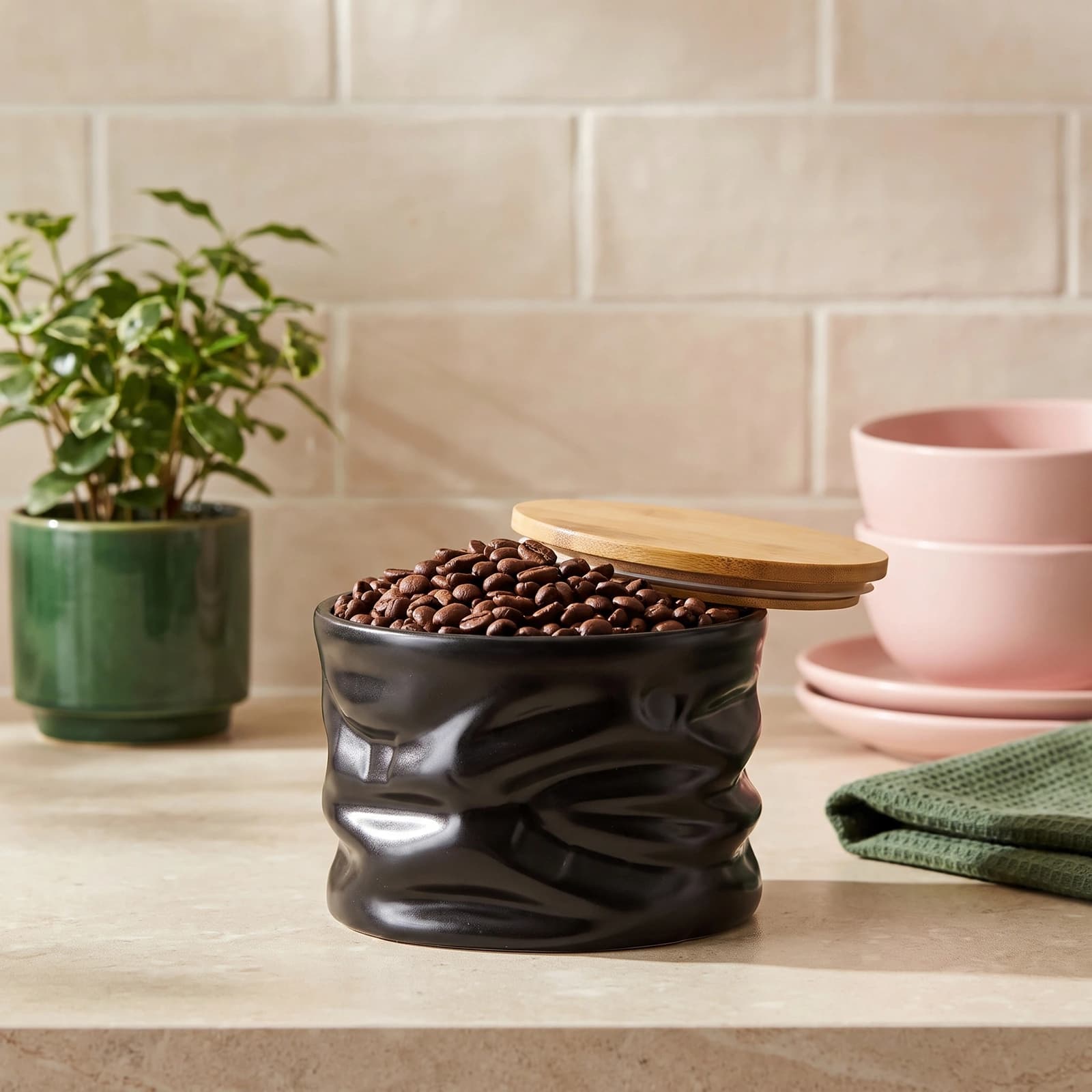 Black crumpled porcelain storage jar with a light wooden lid, filled with coffee beans, on a kitchen counter.