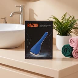 Blue manual razor in black packaging on a light beige bathroom counter next to a white sink and a green plant. - view 1
