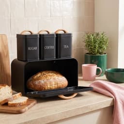 Matte black metal kitchen storage set with three small canisters labeled Sugar, Coffee, Tea, and a large bread box, all with… - view 1
