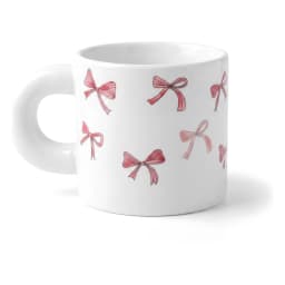 White ceramic mug with a large rounded handle and pink bow patterns, held in a hand, with other mugs in the background. - view 2