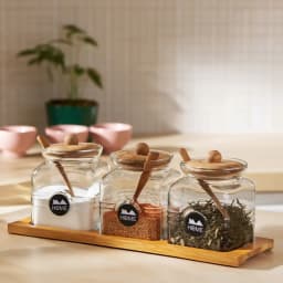 Three clear square glass storage jars with light brown bamboo lids, matching spoons, and a bamboo tray on white background - view 1