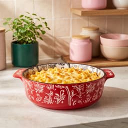 Red granite oval baking dish with white floral patterns, filled with baked macaroni and cheese, on a kitchen counter. - view 1