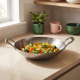 Hammered gray metal wok with two handles, filled with colorful stir-fried chicken and vegetables, on a light kitchen counter. - view 1