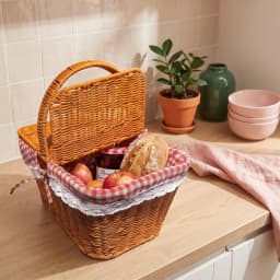 Brown woven picnic basket with red and white gingham lining, filled with grapes, cheese, bread, and a bottle, on a kitchen c… - view 5