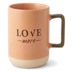 كوباية سيراميك لون خوخي مكتوب عليها 'LOVE more' بالأسود، يد لونها زيتوني وقاعدة بيج فاتح على سطح أبيض. - view 1
