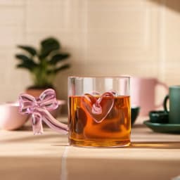 Single clear glass mug with a pink bow-shaped handle and a pink heart decoration on the side, on a white background. - view 1