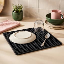 Dark gray square silicone coaster mat with a grid pattern, holding a steaming metal pot on a light brown kitchen counter. - view 3