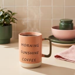 كوب سيراميك برتقالي بعبارة 'MORNING SUNSHINE COFFEE' بالأسود، يد باللون الزيتوني وقاعدة كريمي على رف أبيض. - view 1