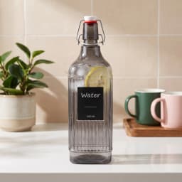 Dark grey ribbed glass bottle with a white and red swing-top stopper, labeled "Water 1000 ML", on a white shelf. - view 1