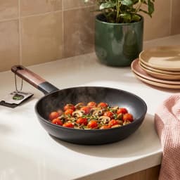 Black granite frying pan with a dark wooden handle, cooking cherry tomatoes and mushrooms with steam rising, on a white kitc… - view 1