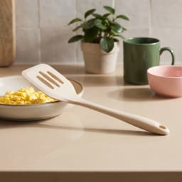 A single beige silicone slotted spatula with a hanging hole, lying diagonally on a white background. - view 1