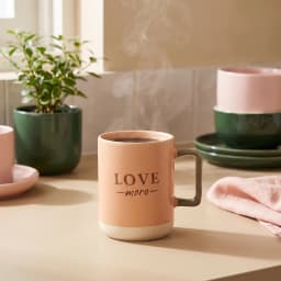 كوباية سيراميك لون خوخي مكتوب عليها 'LOVE more' بالأسود، يد لونها زيتوني وقاعدة بيج فاتح على سطح أبيض. - view 2