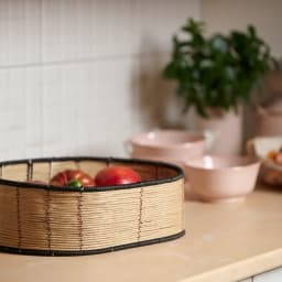 Round shallow storage basket made of light beige woven natural fibers with black coiled edges on a white background. - view 4