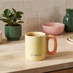 كوب سيراميك أصفر فاتح بيد برتقالي على شكل حلقة وكلمة 'OPTIMISTIC' مكتوبة عليه، معزول على خلفية بيضاء. - view 1