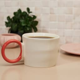 Single cream-colored ceramic mug with a glossy bright red circular handle on a white background. - view 1