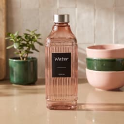 Rose gold ribbed glass water bottle with a silver cap and 'Water 1000 ML' label, on a white shelf. - view 1