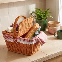 Brown woven picnic basket with red and white gingham lining, filled with grapes, cheese, bread, and a bottle, on a kitchen c… - view 1