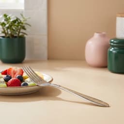 Single silver stainless steel dinner fork with gold accents on handle, next to a fruit plate on a beige counter. - view 1