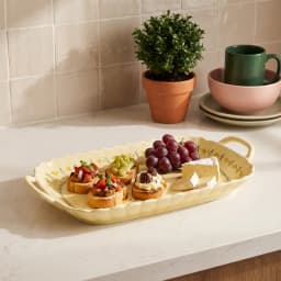 Cream porcelain serving tray with yellow floral design and wavy edges, holding appetizers, grapes, and cheese on a kitchen c… - view 1