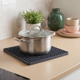Dark gray square silicone coaster mat with a grid pattern, holding a steaming metal pot on a light brown kitchen counter. - view 1
