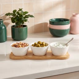 Three white porcelain serving bowls with embossed design on a light brown wood stand, filled with snacks and dip. - view 1